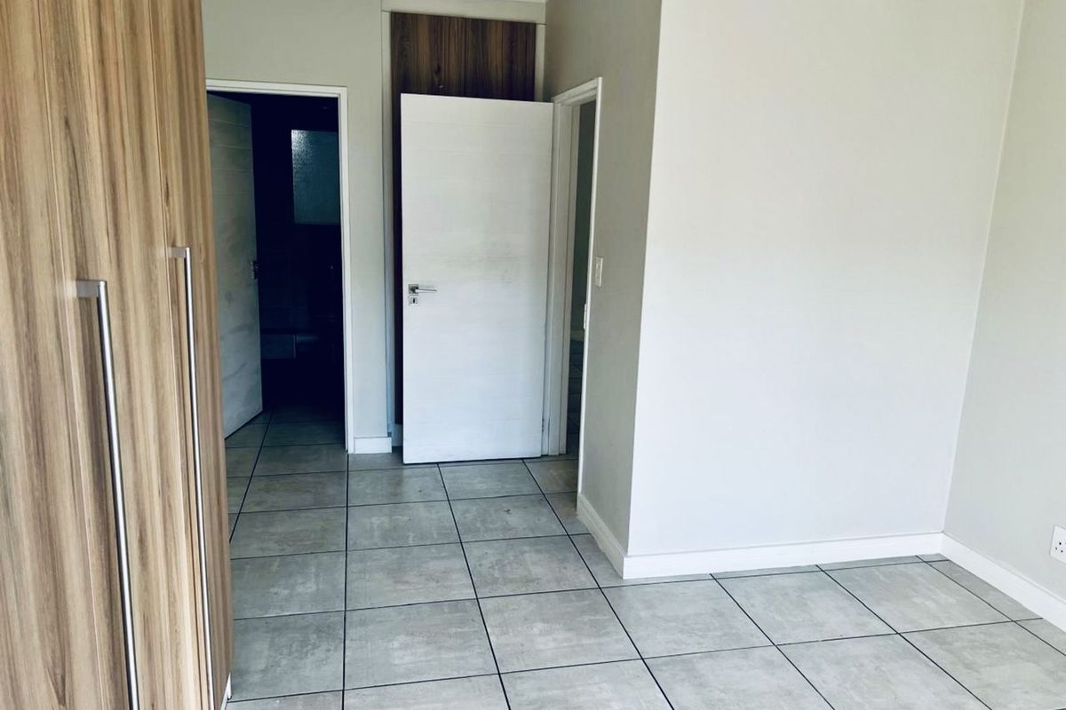 A clean, neutral bedroom featuring large-format floor tiles, built-in wood-grain cupboards, and access to an en-suite bathroom.