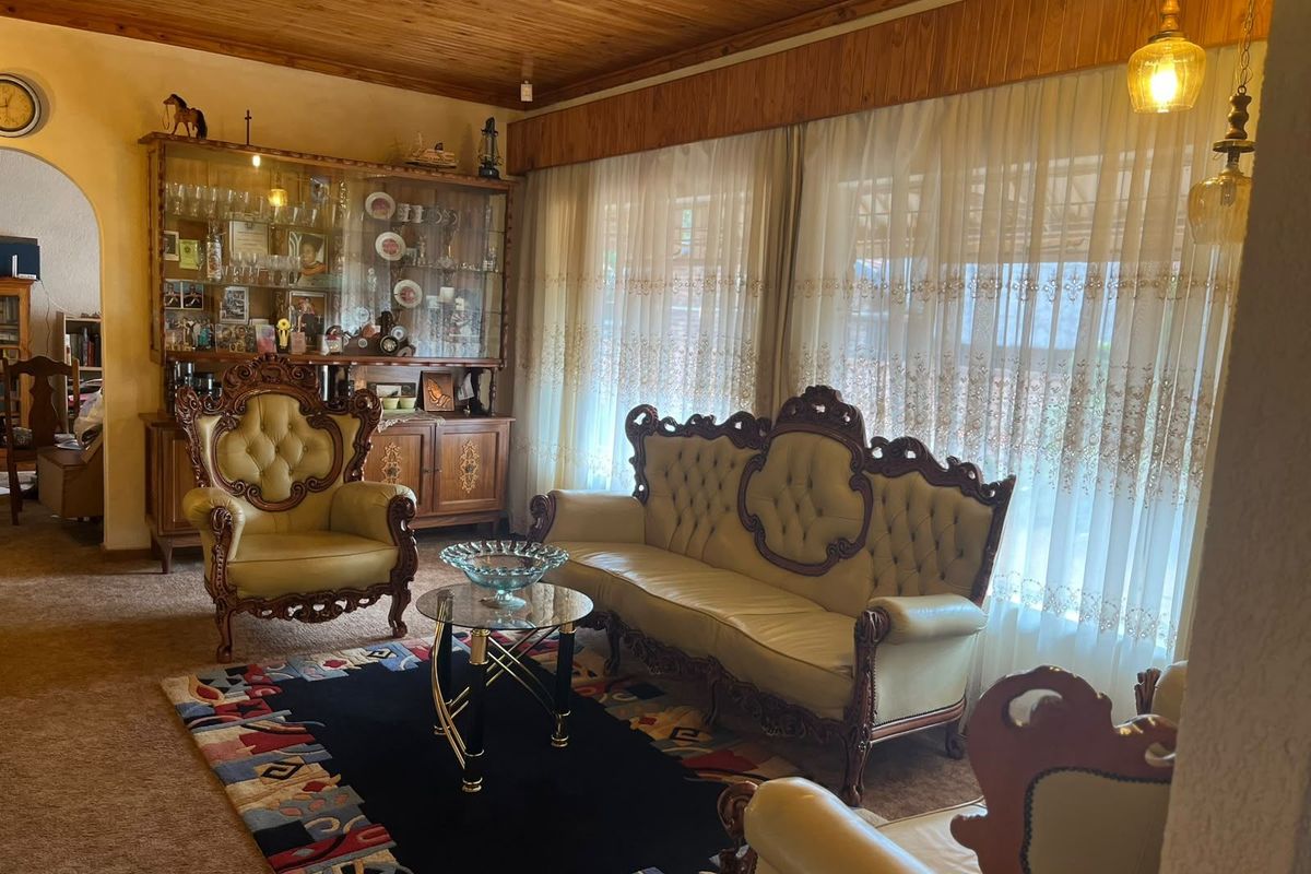 A traditional living room featuring a wood-paneled ceiling, large windows with sheer curtains, and carpeted flooring.