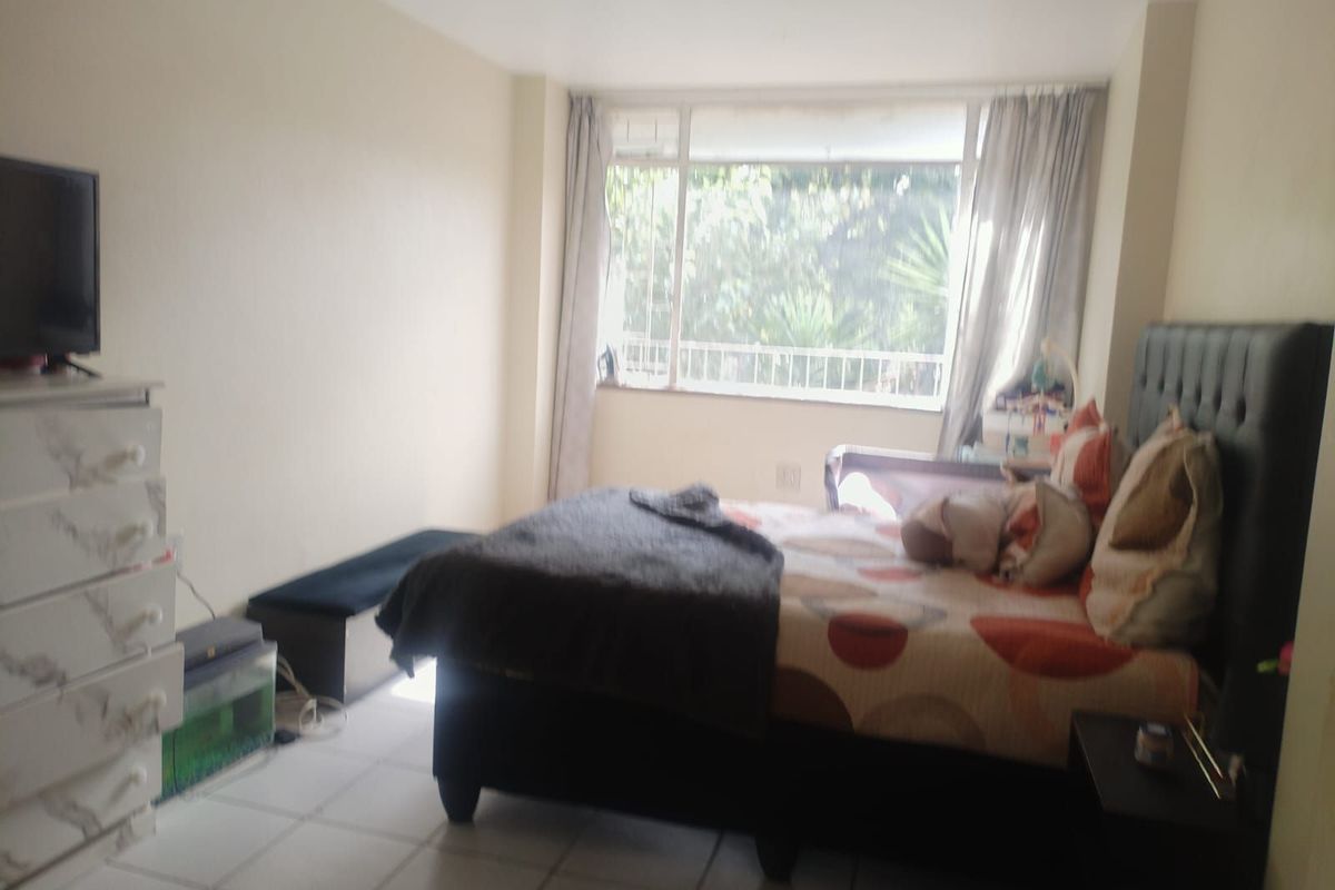 A bedroom featuring tiled flooring and a large window with security bars, providing natural light and a view of the exterior greenery.