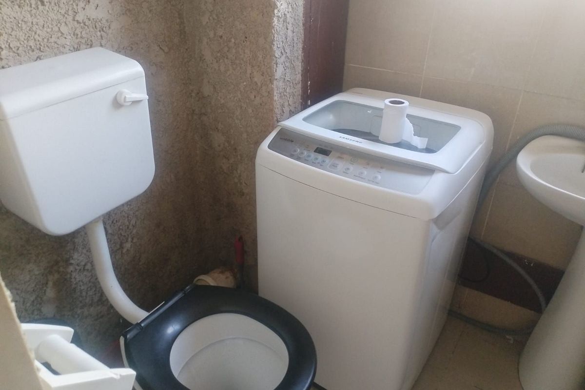 A compact bathroom featuring a toilet, pedestal basin, and space for a washing machine, requiring cosmetic updates and wall finishing.