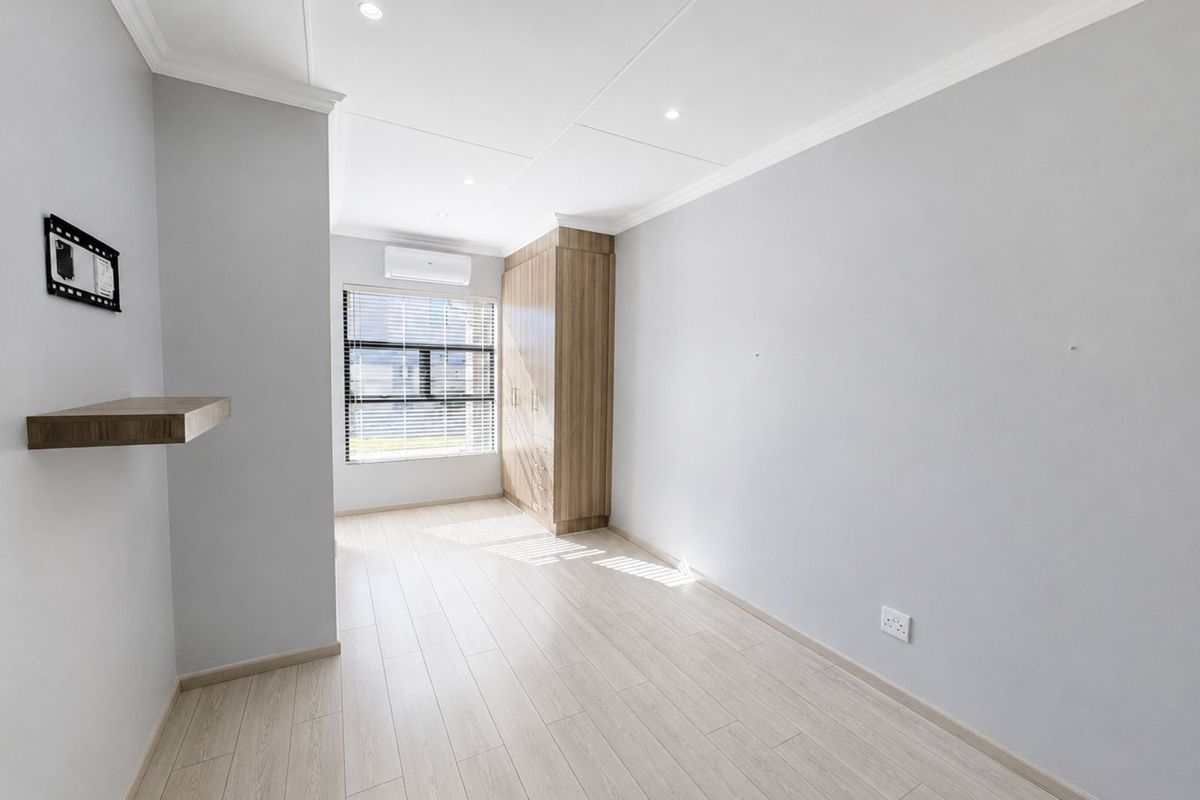 A bright, empty bedroom featuring light wood-look flooring, built-in wooden cabinetry, recessed ceiling lighting, and a wall-mounted air conditioning unit.