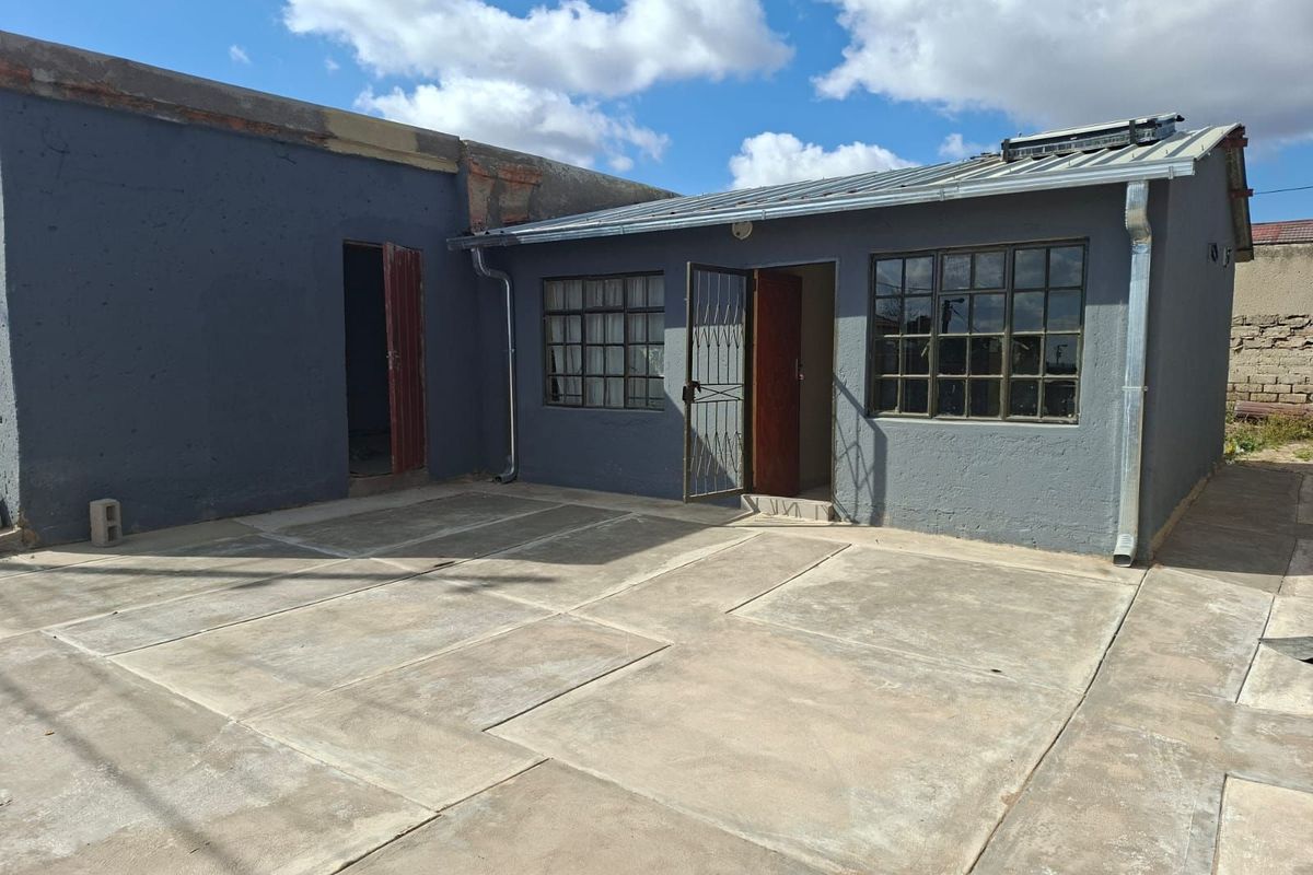 Rear exterior view of the house featuring a paved courtyard, grey painted masonry walls, metal-framed windows, and a solar water heating system on the roof.