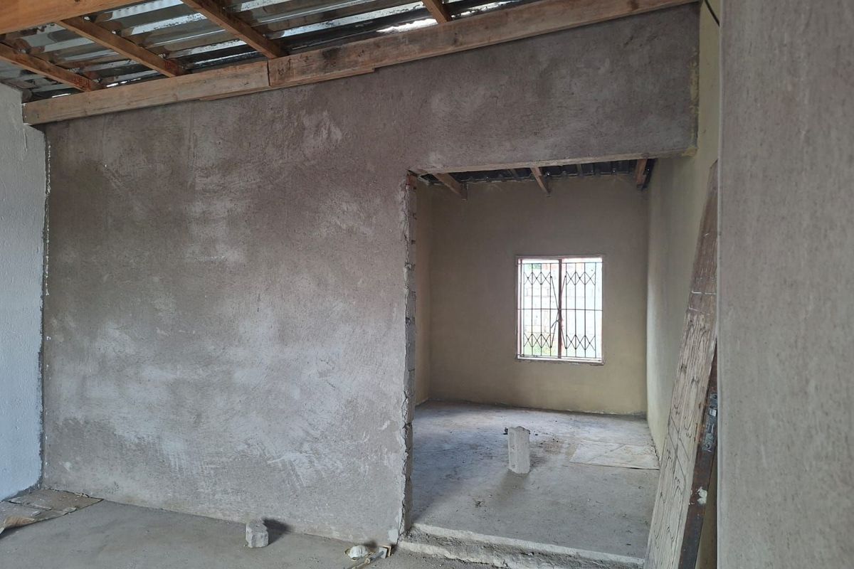 An interior view of a property under construction, featuring raw plastered walls, exposed roof beams, and a window with security bars.