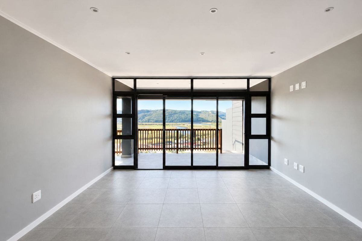 A bright, modern, and empty living room featuring large sliding glass doors that open onto a balcony with scenic landscape views.