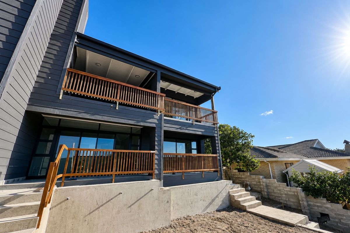 A modern two-story exterior rear view featuring dark timber cladding, expansive glass sliding doors, and wooden balcony railings overlooking an unfinished garden area.
