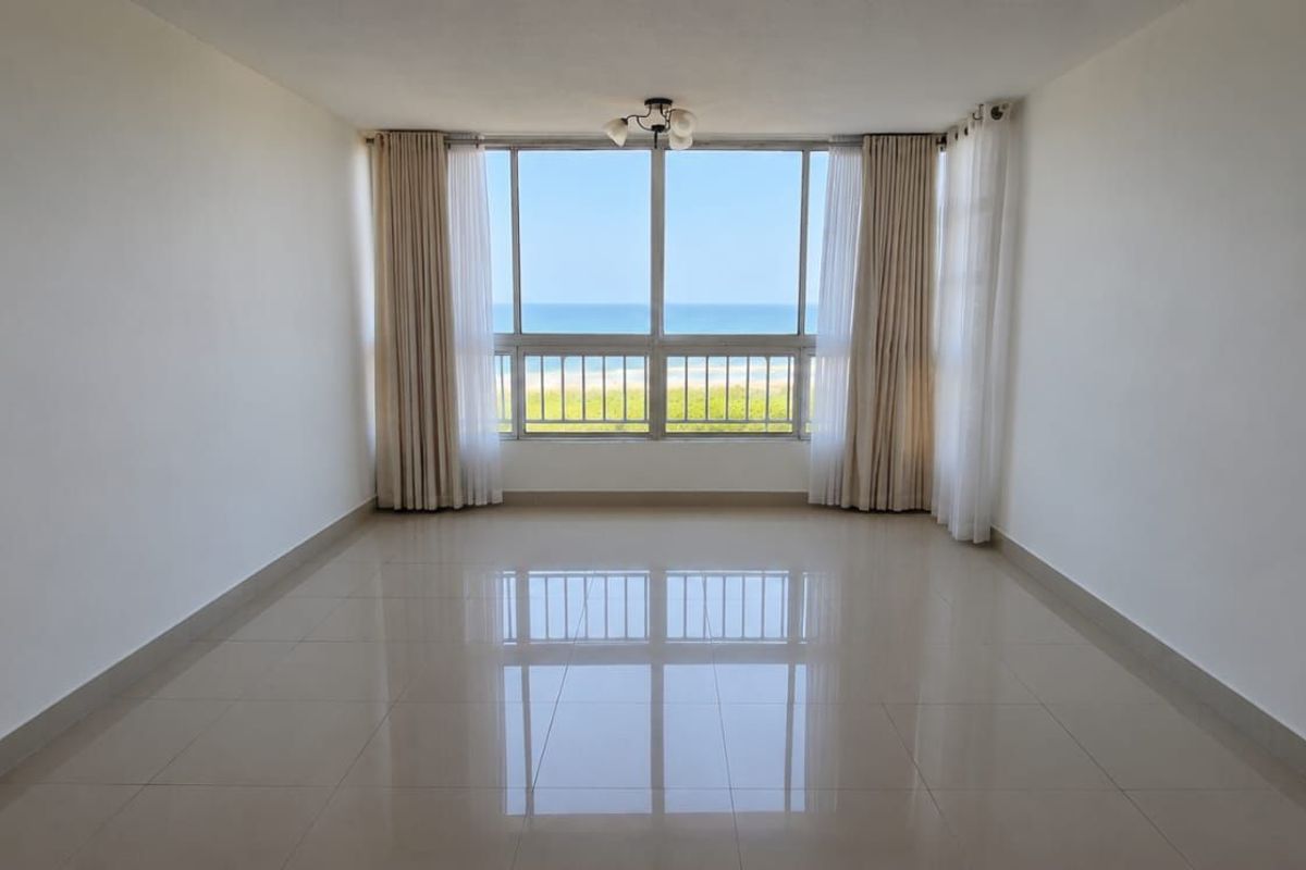 A bright, empty living room featuring large windows with direct ocean views and polished tile flooring.
