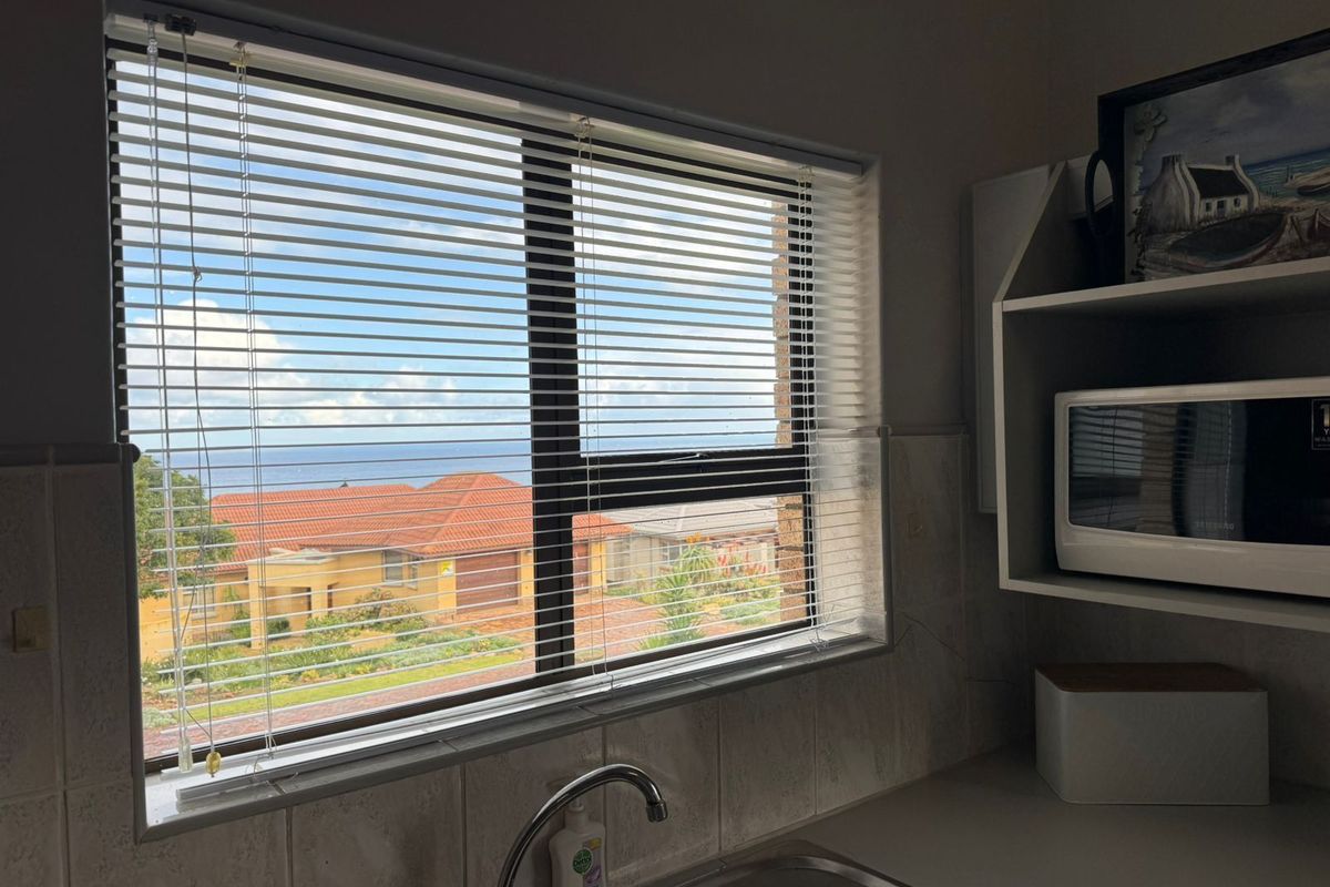 A kitchen sink area featuring a stainless steel basin, tiled splashback, and a window with horizontal blinds overlooking a residential neighborhood and distant sea view.
