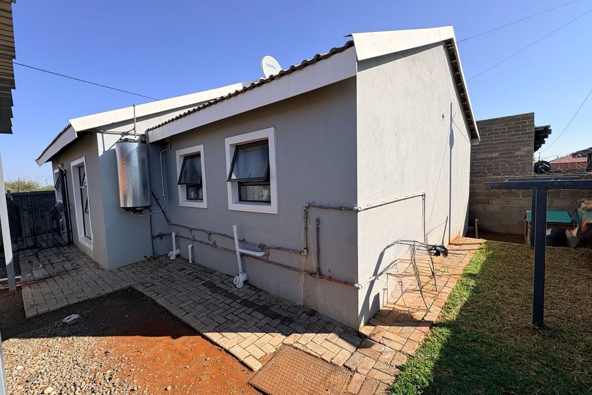 Rear exterior view of the house featuring paved walkways, external geyser unit, and exposed plumbing infrastructure along the side wall.