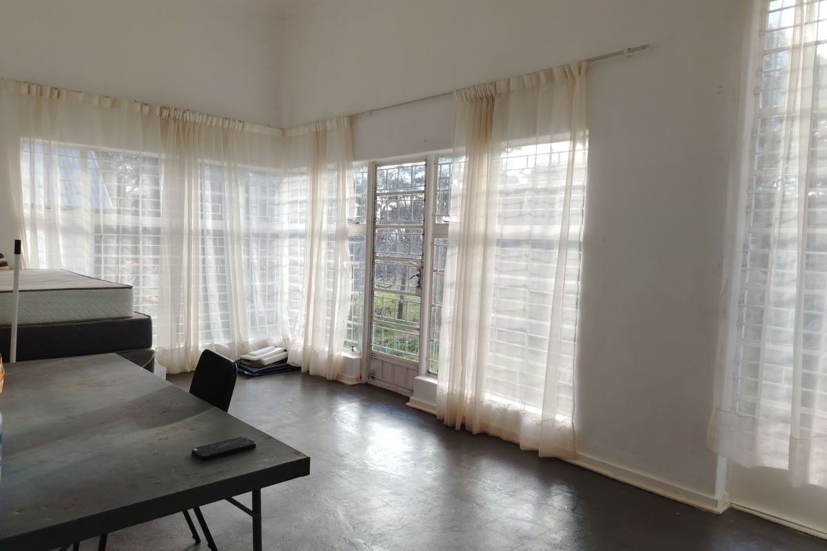 A bright, spacious living area featuring large windows with security bars, sheer curtains, and dark painted flooring, offering ample natural light.