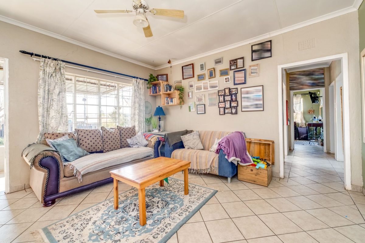A living room featuring tiled flooring, a ceiling fan, and large windows with security bars, providing access to the adjacent hallway.