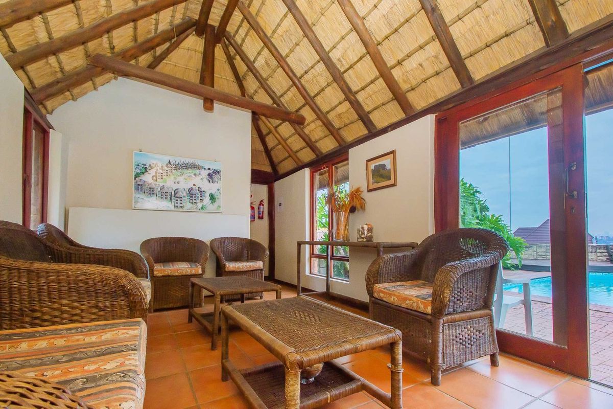 A cozy living area featuring a high vaulted thatch ceiling, terracotta floor tiles, and large glass doors opening onto a pool deck.