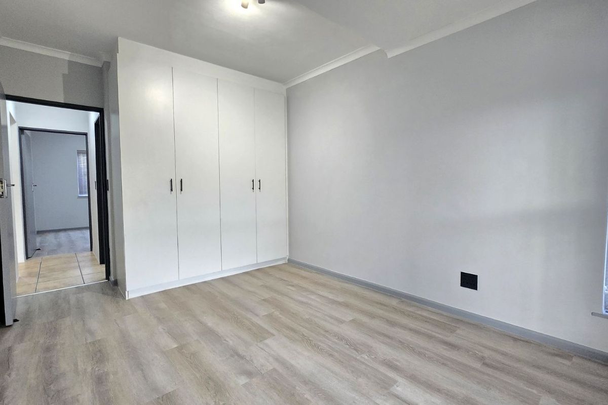 A bright, empty bedroom featuring modern light wood-look flooring, built-in white cupboards, and a large window with blinds.