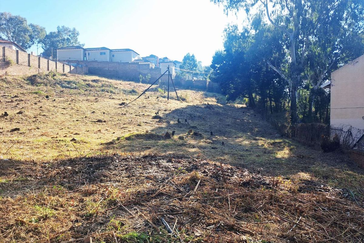 A sloped, undeveloped residential plot featuring natural terrain, mature trees, and perimeter boundary walls in a suburban setting.