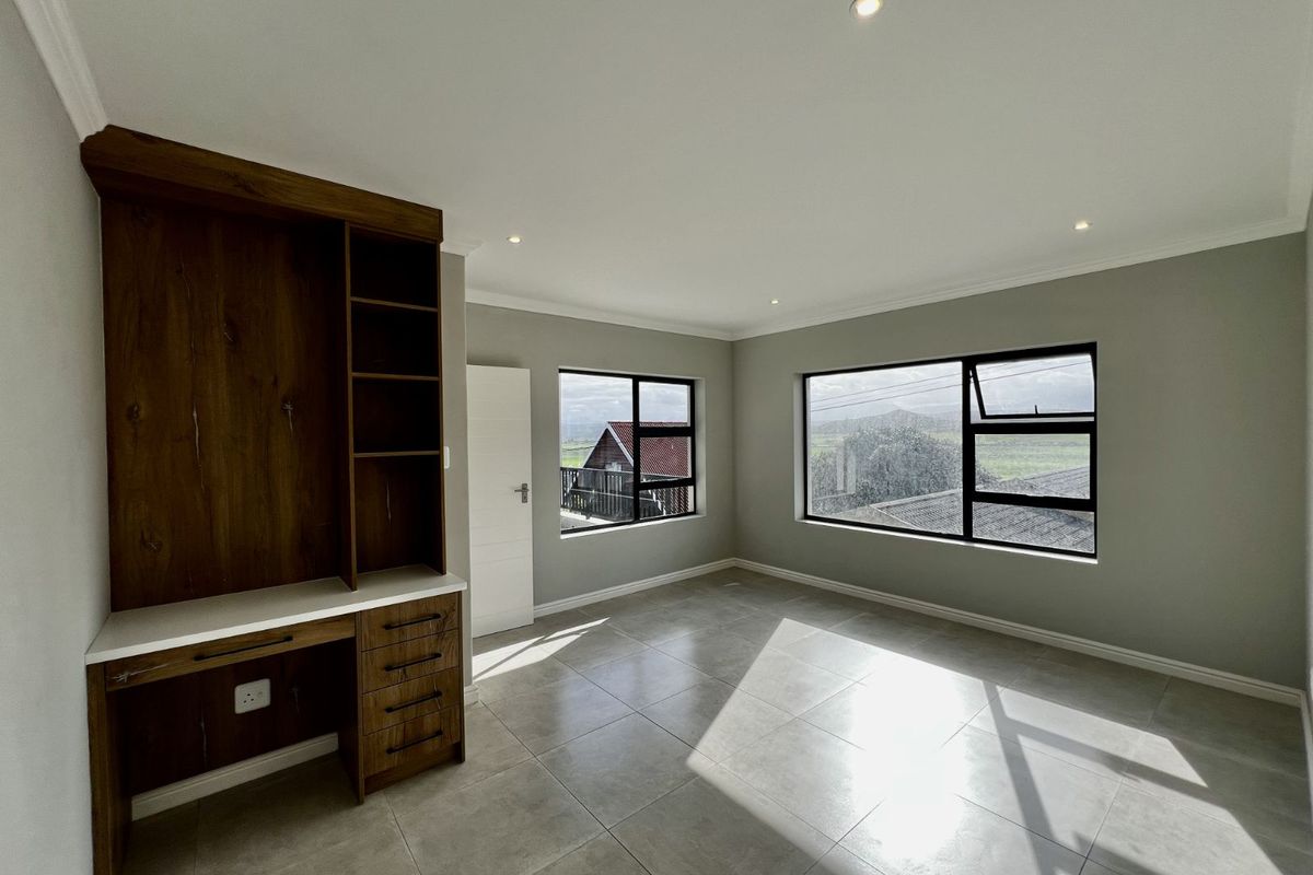 A bright, modern bedroom featuring large windows, neutral tiled flooring, recessed ceiling lighting, and a built-in wooden study desk with shelving.