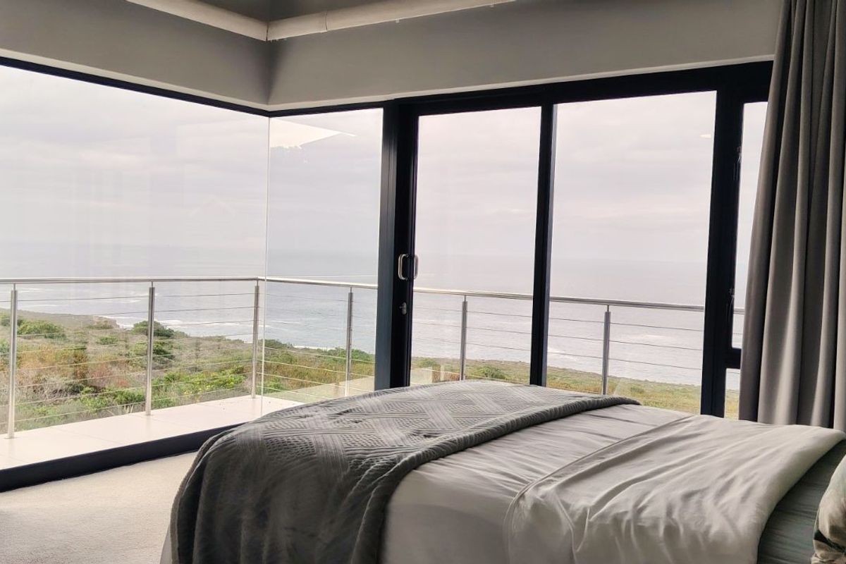 A modern bedroom featuring expansive floor-to-ceiling corner windows and sliding glass doors that open onto a private balcony with panoramic ocean views.
