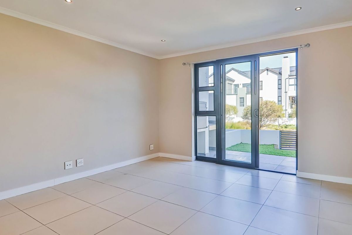 A bright, empty living room featuring neutral tiled flooring, recessed ceiling lighting, and large sliding glass doors providing access to an outdoor patio area.