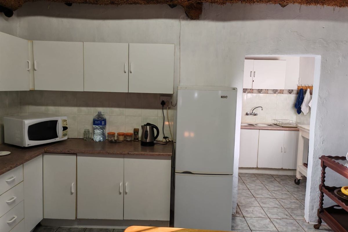 A dated kitchen featuring white cabinetry, laminate countertops, and a tiled splashback, with an adjoining scullery area visible through the doorway.