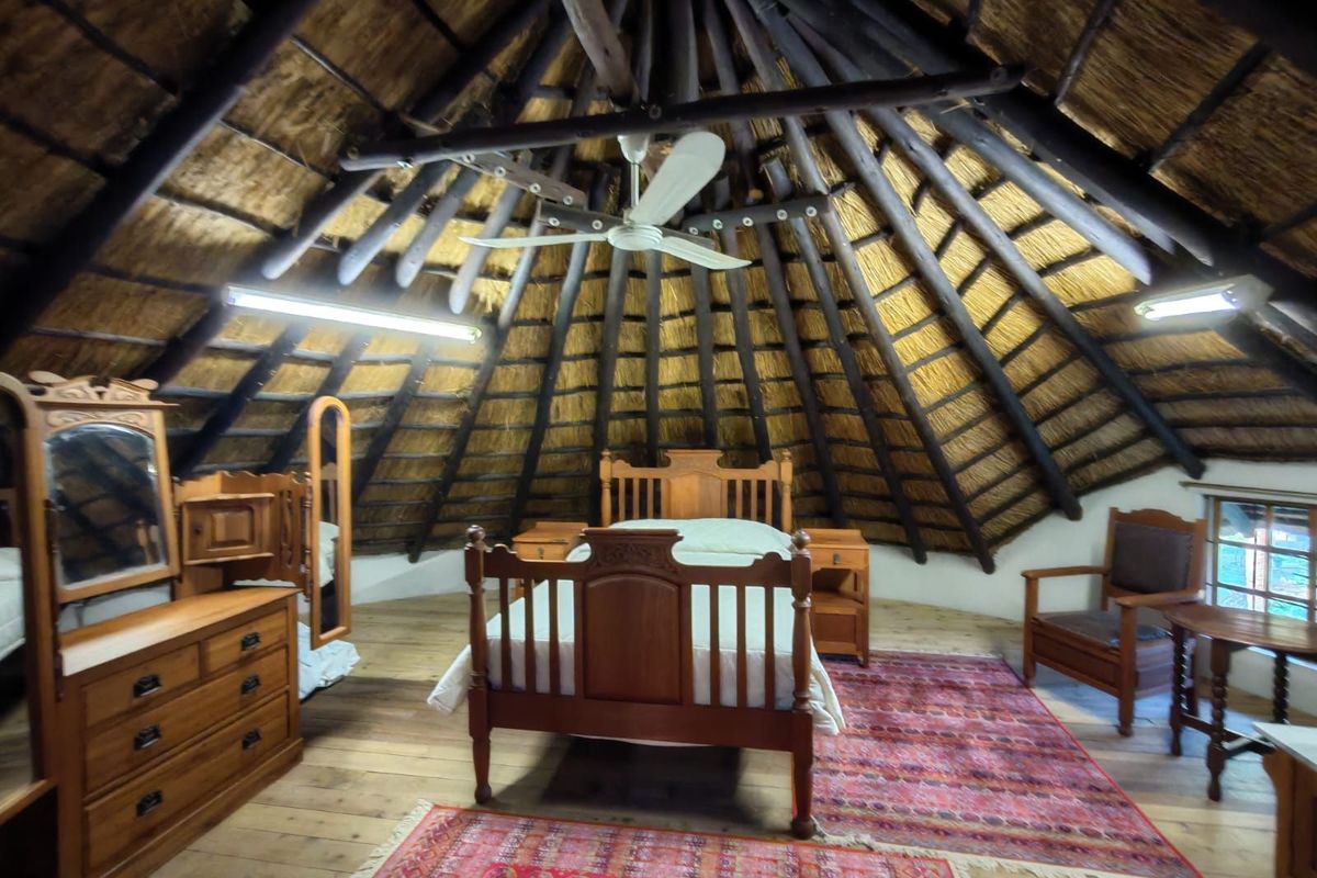 A unique bedroom featuring a traditional thatched roof with exposed timber rafters, wooden flooring, and a central ceiling fan.