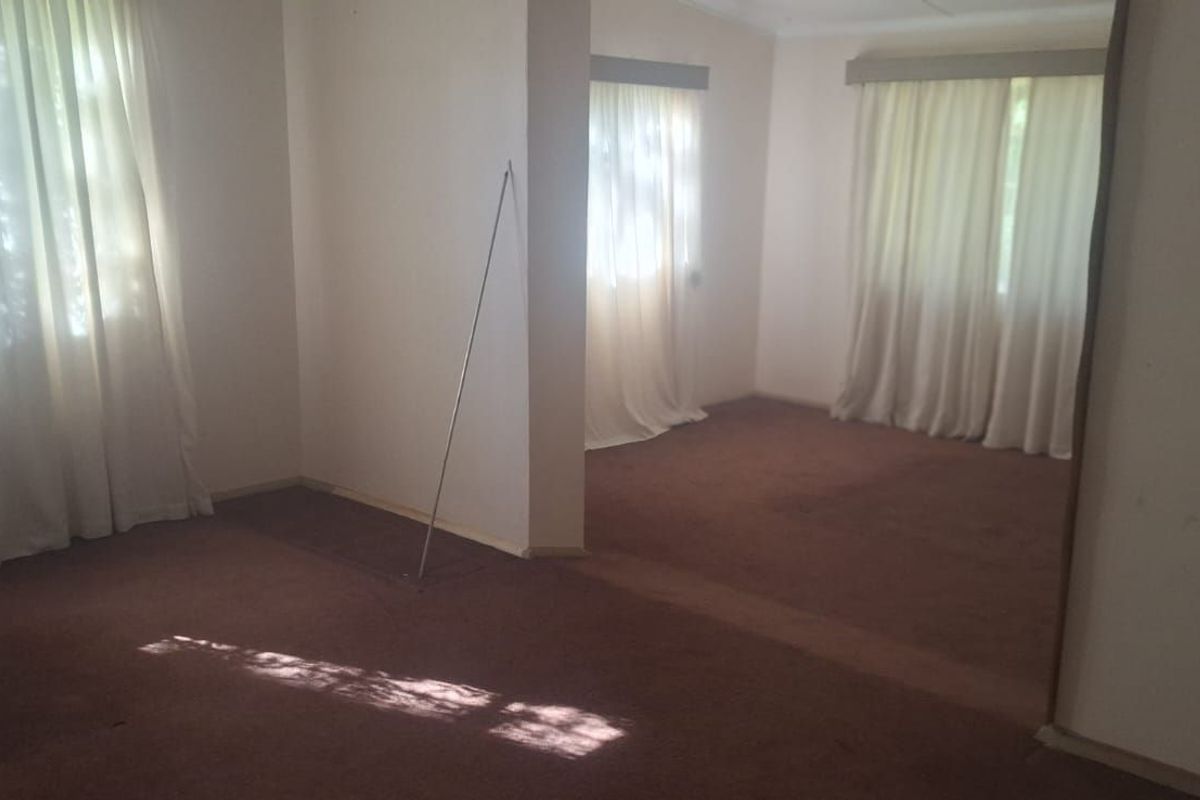 A spacious, empty living area featuring neutral walls, carpeted flooring, and large windows with curtains, offering a blank canvas for potential renovation.