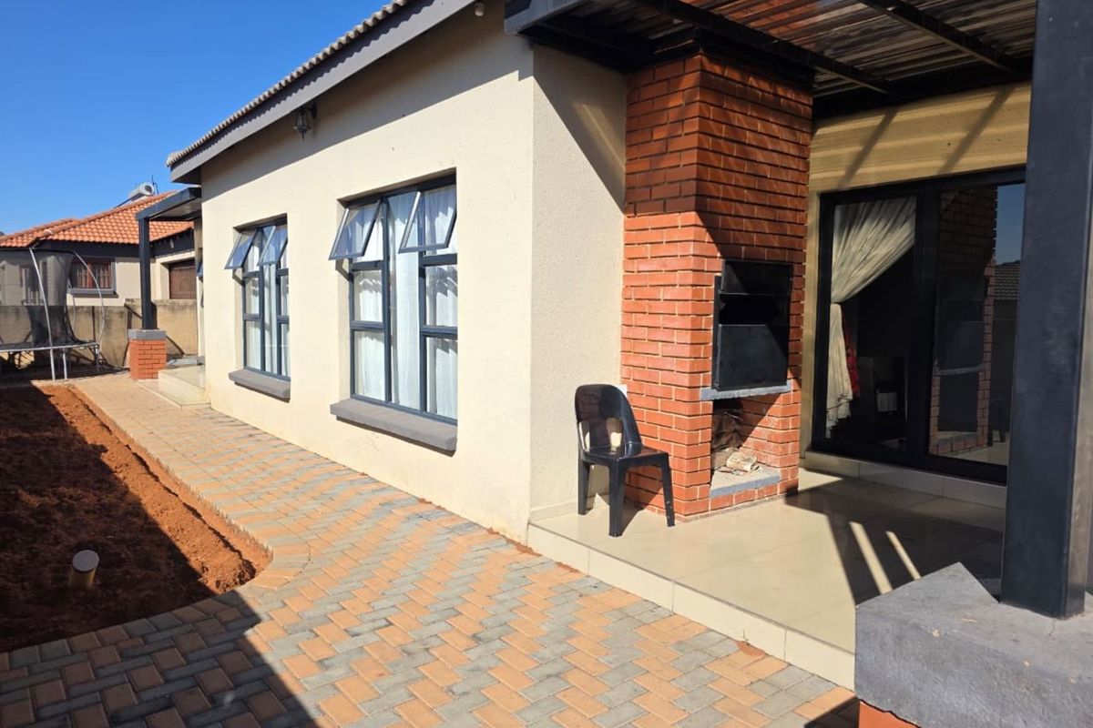 Rear exterior view featuring a paved patio area with a built-in brick braai, tiled flooring, and aluminium window frames.