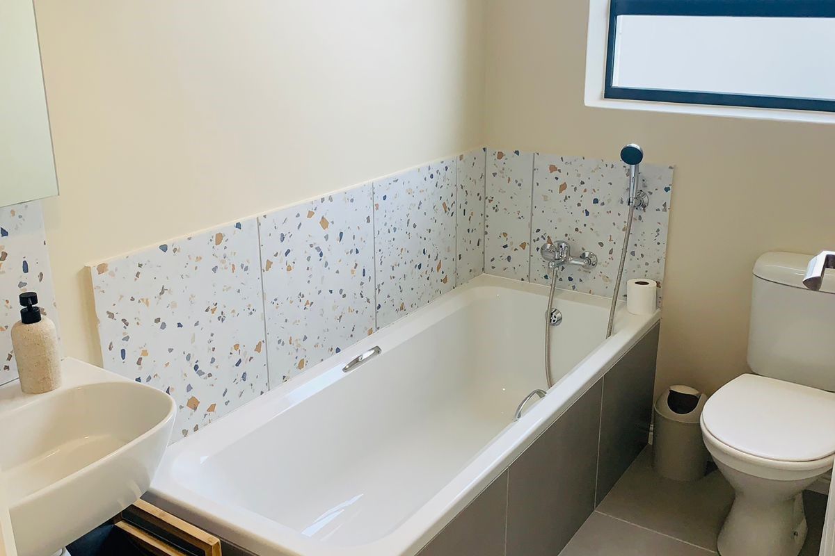 A clean, modern bathroom featuring a bathtub with terrazzo-style splashback tiles, a wall-mounted basin, and a toilet.