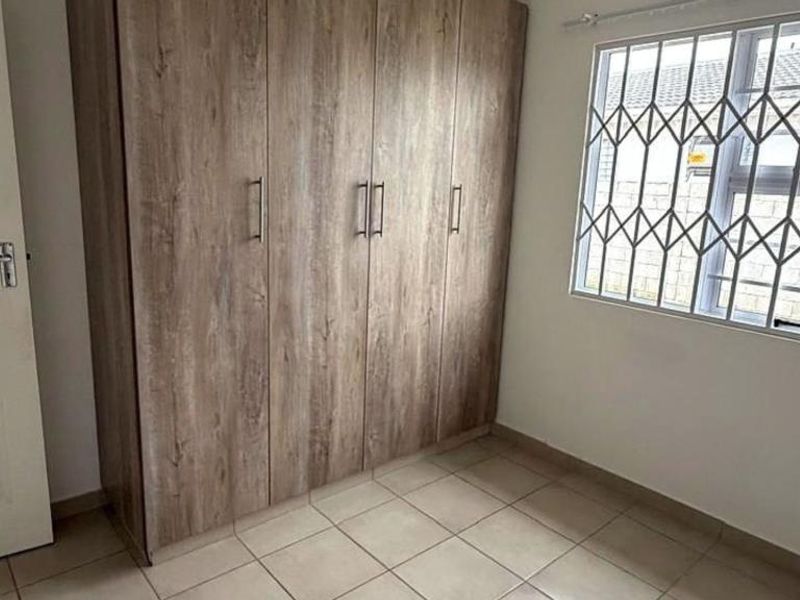 A clean, empty bedroom featuring built-in wooden wardrobes, tiled flooring, and a window fitted with a security gate.