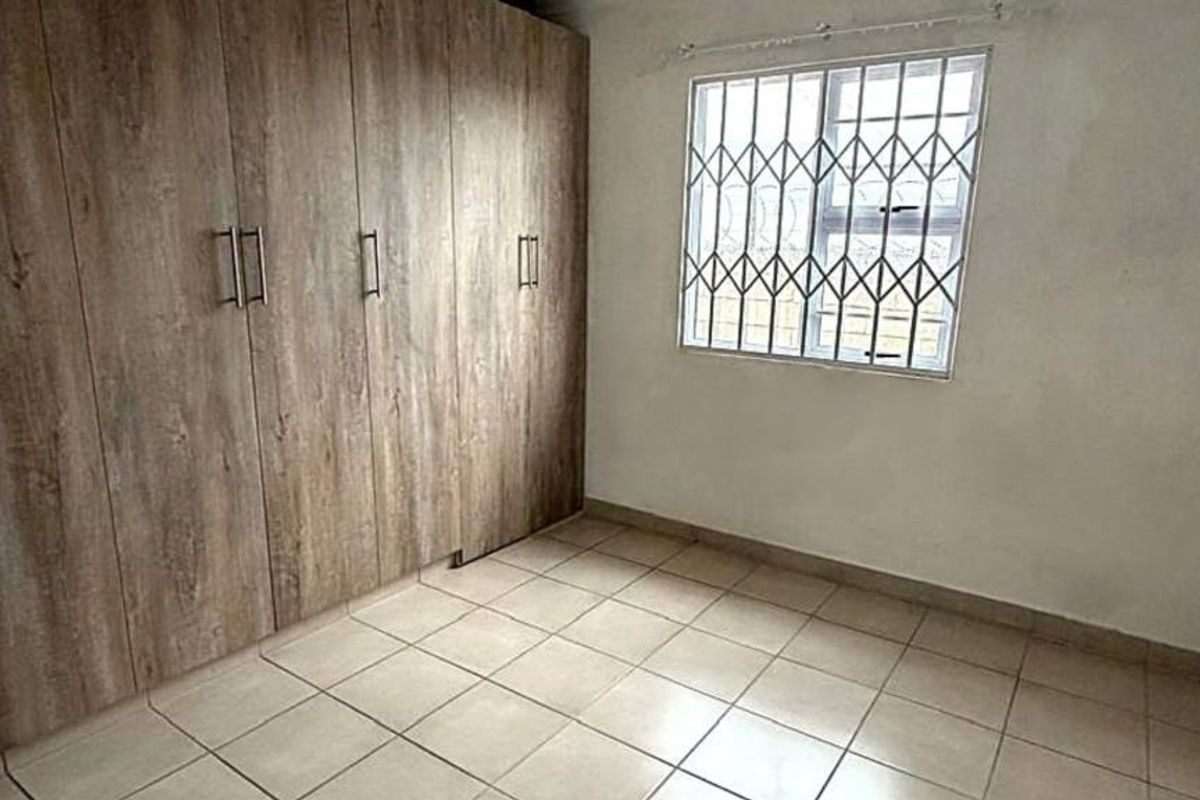 A functional bedroom featuring tiled flooring, built-in wooden cupboards, and a window fitted with security bars.