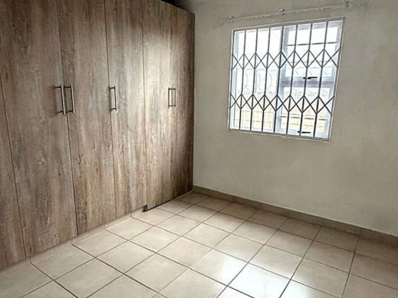 A functional bedroom featuring tiled flooring, built-in wooden cupboards, and a window fitted with security bars.