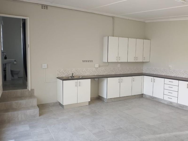 A clean, empty kitchen featuring white built-in cabinetry, dark laminate countertops, tiled backsplash, and tiled flooring, with an adjacent doorway leading to a bathroom.