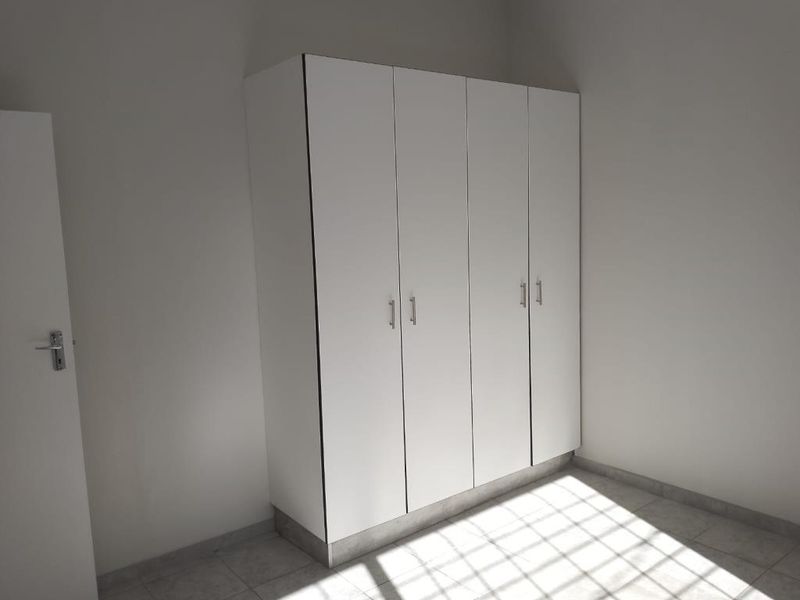 A bright, empty bedroom featuring a clean, white built-in wardrobe with modern handles and tiled flooring.