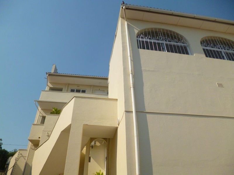 A low-angle view of the apartment building exterior, featuring cream-colored walls, arched windows with security bars, and an external balcony structure.