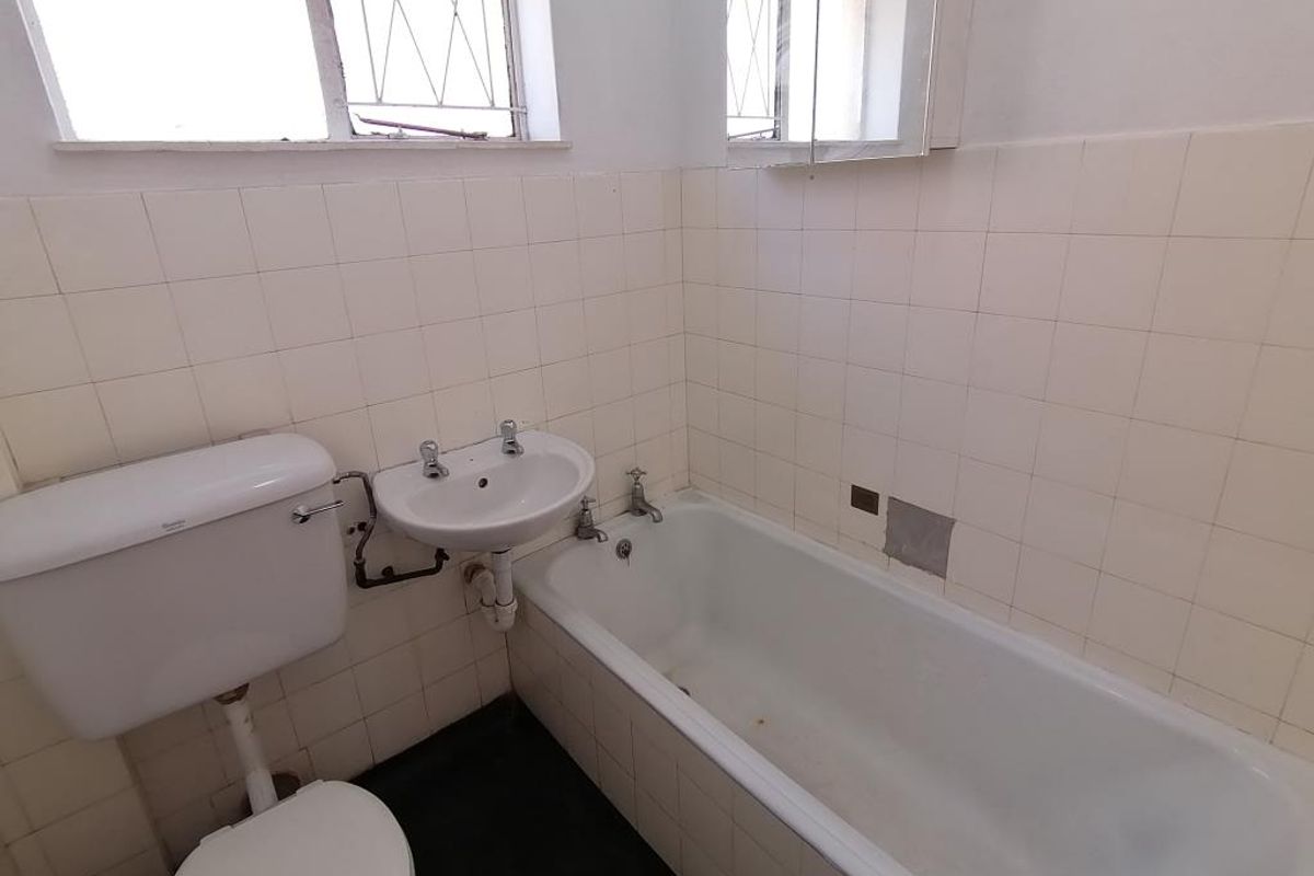 A dated bathroom featuring a standard bathtub, wall-mounted basin, and toilet, with partially tiled walls and a window for natural light.