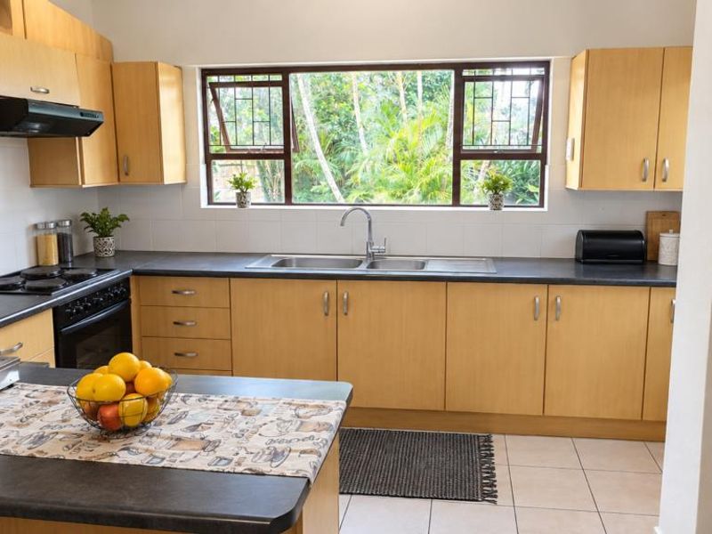 A functional kitchen featuring light wood cabinetry, dark laminate countertops, a double sink, and built-in cooking appliances with a view of the garden.