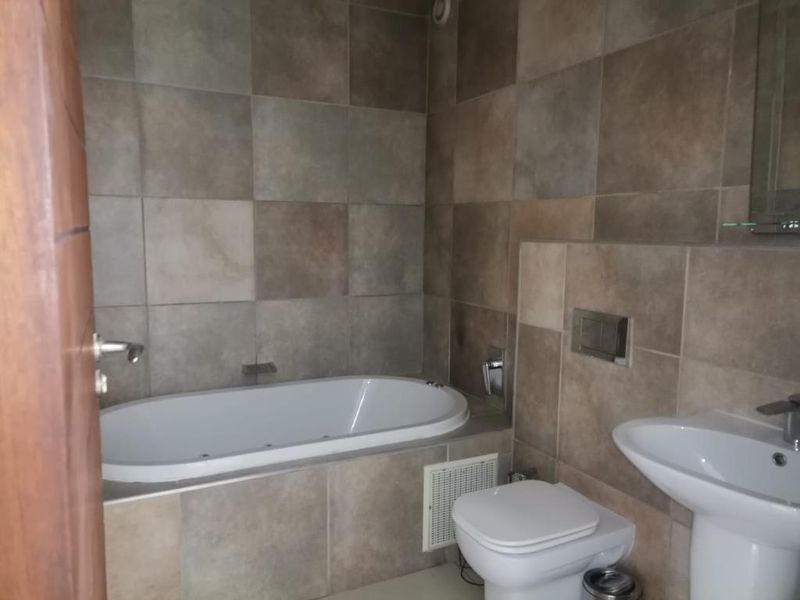 A functional bathroom featuring a built-in bathtub, pedestal sink, and wall-mounted toilet with neutral ceramic wall tiling throughout.