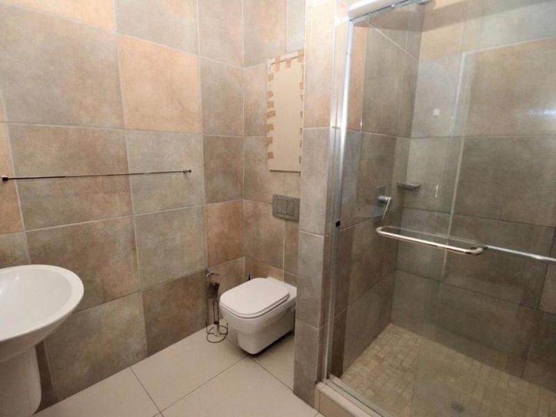 A modern bathroom featuring a pedestal basin, wall-hung toilet with concealed cistern, and a glass-enclosed shower with neutral wall and floor tiling.