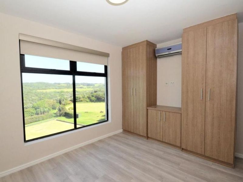 A bright, empty bedroom featuring modern light wood built-in cupboards, laminate flooring, an air conditioning unit, and a large window with scenic views.