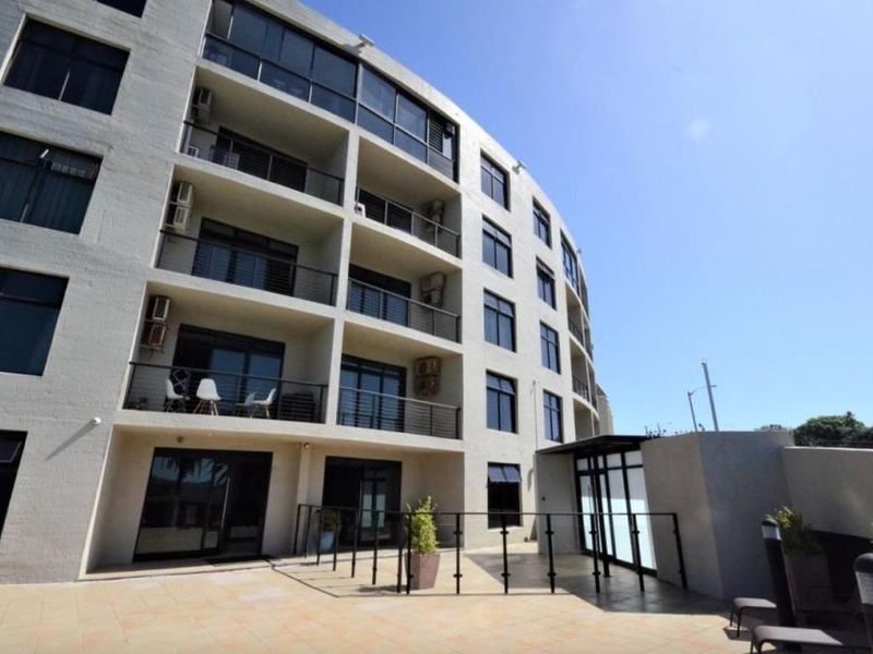 A view of the rear facade of a modern apartment building featuring multiple levels of balconies with glass balustrades and a paved patio area.