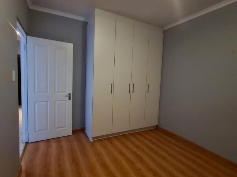 A clean, empty bedroom featuring light grey walls, warm-toned laminate flooring, and white built-in cupboards with modern finishes.