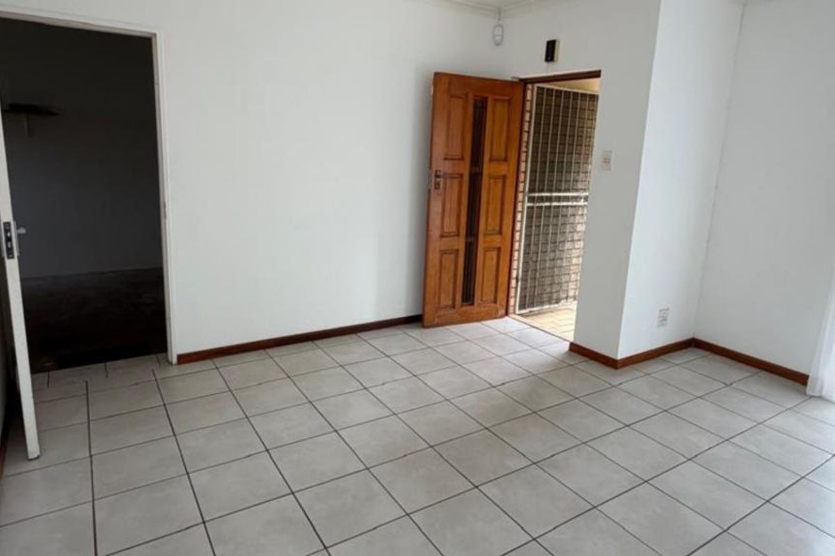 An empty living area featuring neutral ceramic floor tiles, a timber entrance door with security gate, and access to an adjacent room.