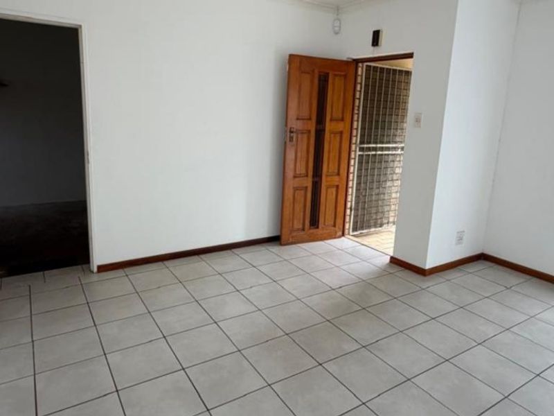 An empty living area featuring neutral ceramic floor tiles, a timber entrance door with security gate, and access to an adjacent room.