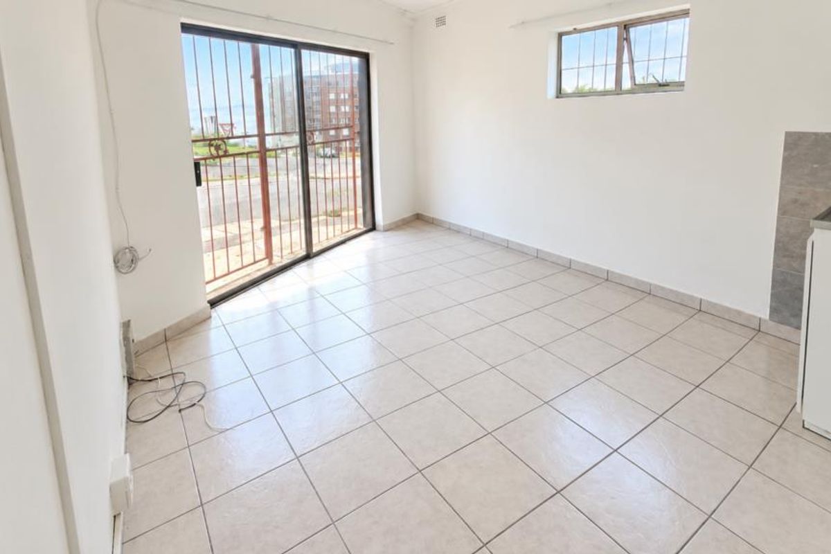 An empty open-plan living area featuring tiled flooring, a sliding glass door with security gate, and a basic kitchen sink area.