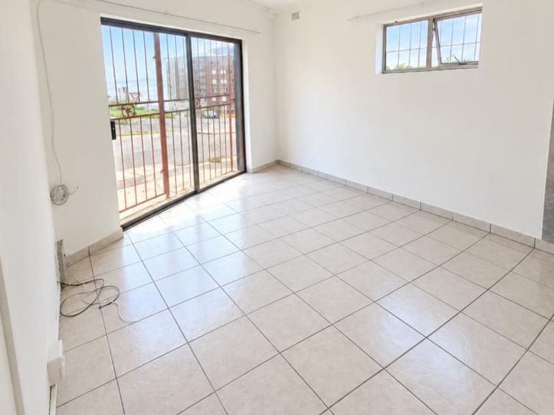 An empty open-plan living area featuring tiled flooring, a sliding glass door with security gate, and a basic kitchen sink area.