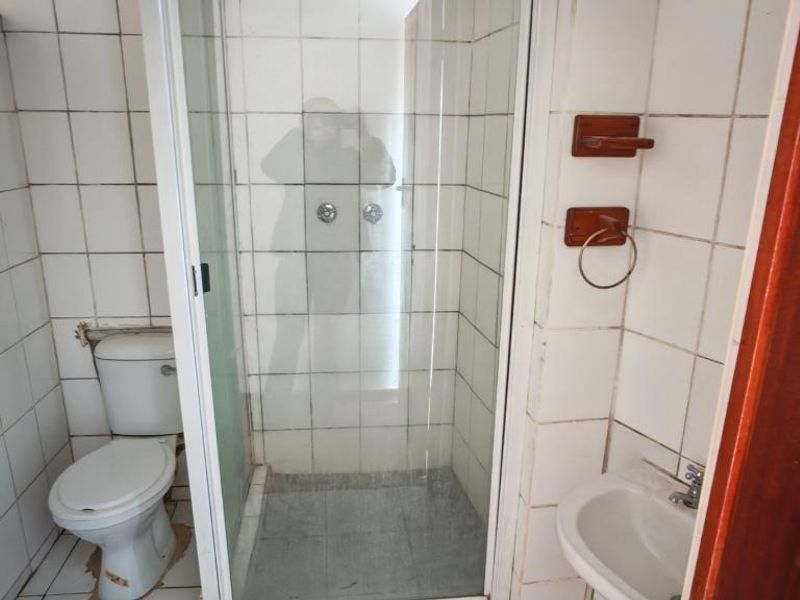 A compact bathroom featuring a shower enclosure, toilet, and wall-mounted basin with dated white wall tiling and floor tiles requiring maintenance.