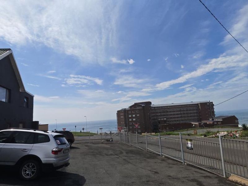 Exterior view of the apartment complex frontage featuring an asphalt parking area and metal perimeter fencing with a coastal backdrop.