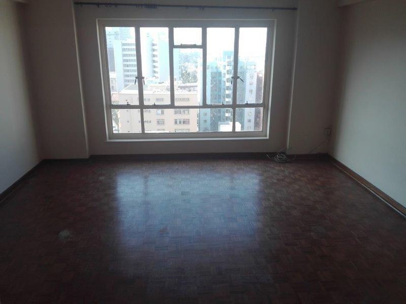 An empty living room featuring classic parquet flooring and a large window with urban city views.