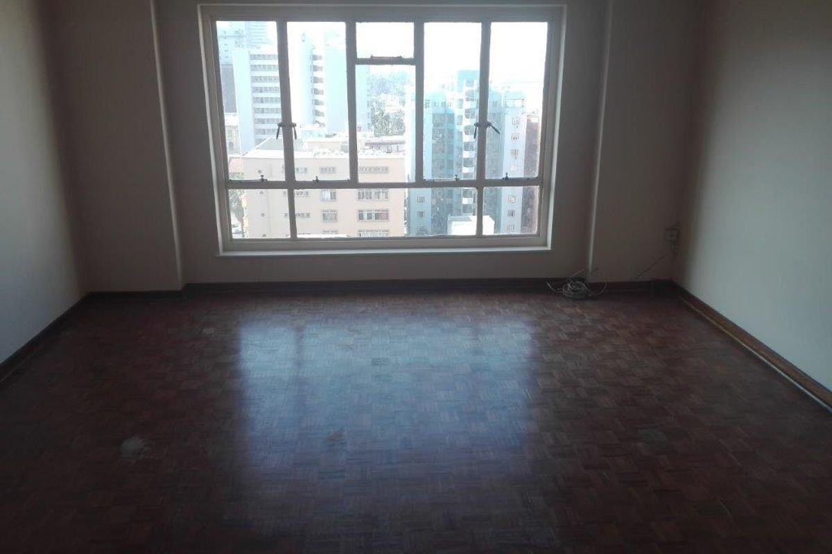 An empty living room featuring classic parquet flooring and a large window with urban city views.