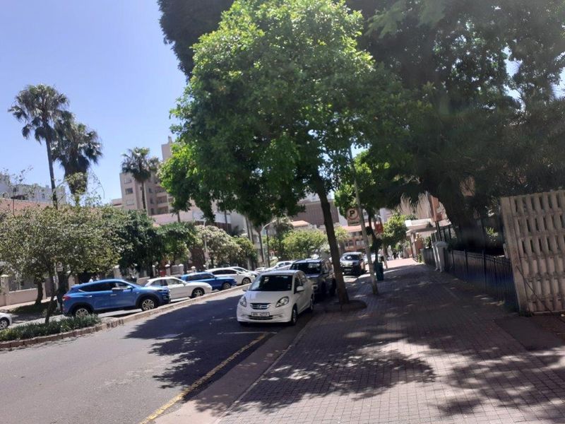 A street-level view showing the paved sidewalk, mature trees, and perimeter fencing along the property boundary.
