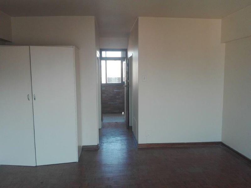 A vacant hallway featuring parquet flooring and a built-in cupboard, leading towards a windowed area.