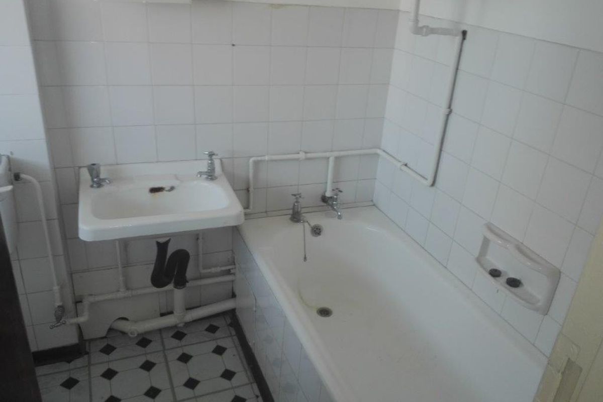 A dated bathroom featuring a wall-mounted basin, bathtub, and exposed plumbing with original ceramic tiling throughout.