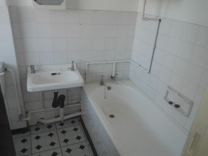 A dated bathroom featuring a wall-mounted basin, bathtub, and exposed plumbing with original ceramic tiling throughout.